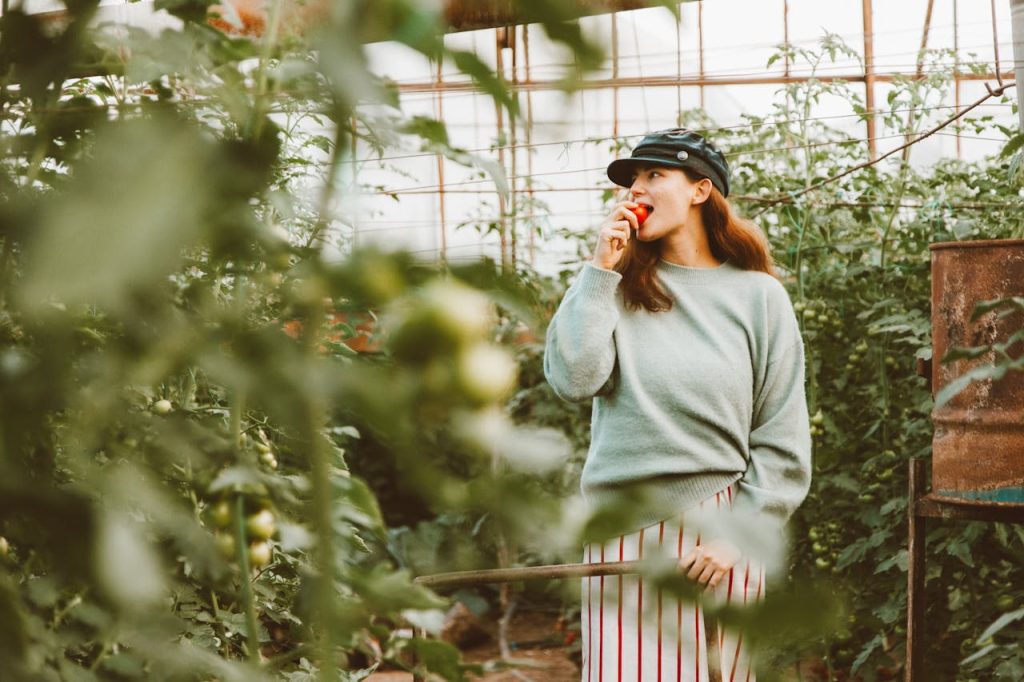Adult woman enjoying a fresh tomato in a lush greenhouse setting.
